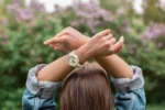 Person seen from behind with brown hair, wearing a denim jacket and raising crossed arms above the head, wearing a white wristwatch with a light green dial, in an outdoor setting with a blurred background of greenery and lilac flowers, symbolizing reflection on the timing of spiritual awakening.