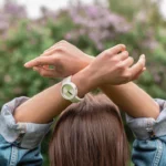 Person seen from behind with brown hair, wearing a denim jacket and raising crossed arms above the head, wearing a white wristwatch with a light green dial, in an outdoor setting with a blurred background of greenery and lilac flowers, symbolizing reflection on the timing of spiritual awakening.