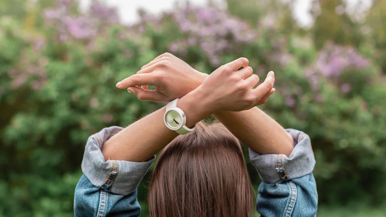 Person seen from behind with brown hair, wearing a denim jacket and raising crossed arms above the head, wearing a white wristwatch with a light green dial, in an outdoor setting with a blurred background of greenery and lilac flowers, symbolizing reflection on the timing of spiritual awakening.