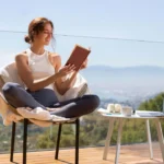 Smiling young woman wearing a white tank top and gray leggings, sitting cross-legged on a cozy armchair, reading a book on a glass balcony with a view of mountains, city, and blue sky — conveying a sense of peace and well-being in the context of spiritual awakening.