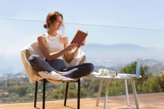 Smiling young woman wearing a white tank top and gray leggings, sitting cross-legged on a cozy armchair, reading a book on a glass balcony with a view of mountains, city, and blue sky — conveying a sense of peace and well-being in the context of spiritual awakening.