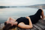 Young woman lying on her back on a wooden deck, wearing a black dress and light sandals, gazing peacefully at the sky. A calm lake and mountains appear in the background, conveying tranquility and introspection associated with spiritual awakening.