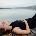 Young woman lying on her back on a wooden deck, wearing a black dress and light sandals, gazing peacefully at the sky. A calm lake and mountains appear in the background, conveying tranquility and introspection associated with spiritual awakening.