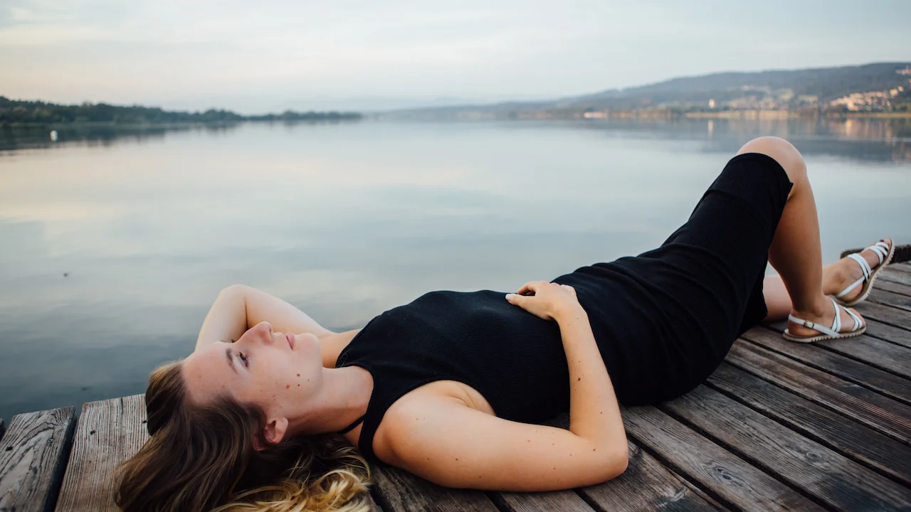 Young woman lying on her back on a wooden deck, wearing a black dress and light sandals, gazing peacefully at the sky. A calm lake and mountains appear in the background, conveying tranquility and introspection associated with spiritual awakening.