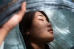 Woman with closed eyes and a serene expression, partially covered by a light blue translucent fabric with floral patterns, softly illuminated by filtered natural light — symbolizing tranquility and expanded consciousness after the awakening.