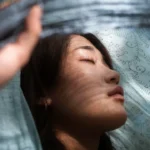 Woman with closed eyes and a serene expression, partially covered by a light blue translucent fabric with floral patterns, softly illuminated by filtered natural light — symbolizing tranquility and expanded consciousness after the awakening.