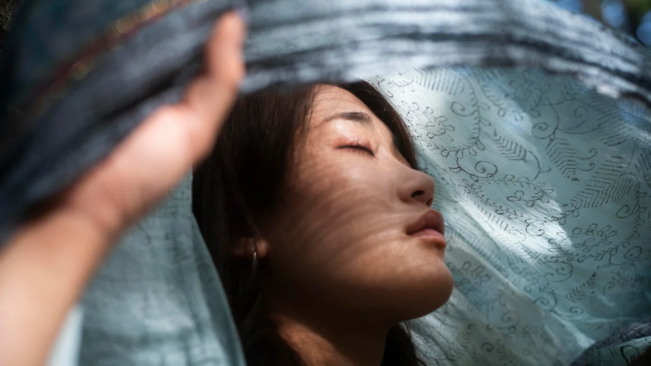 Woman with closed eyes and a serene expression, partially covered by a light blue translucent fabric with floral patterns, softly illuminated by filtered natural light — symbolizing tranquility and expanded consciousness after the awakening.